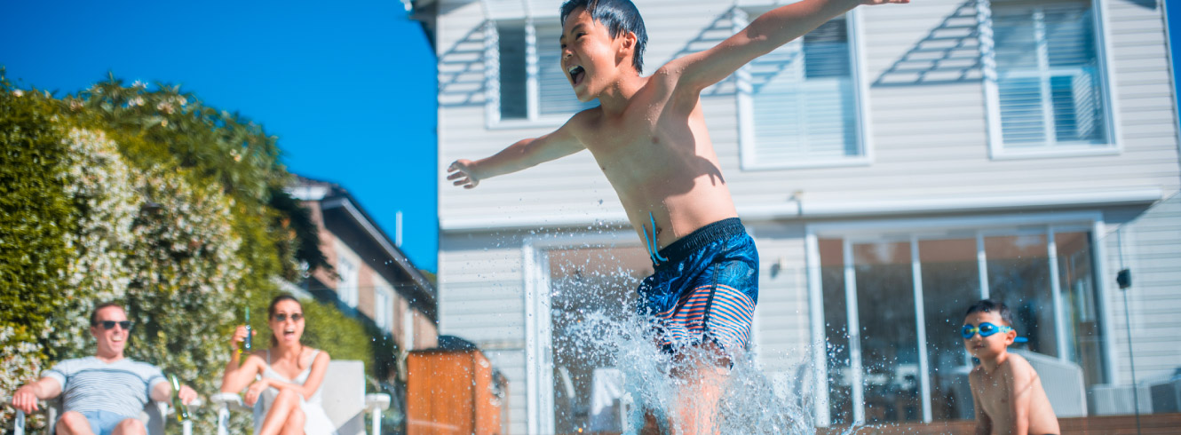 A young boy jumping into a pool feet first. In the background, his family is watching him.