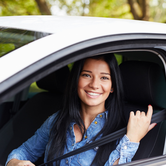 Young woman pulling her seatbelt on inside of a car, she is smiling at the camera.